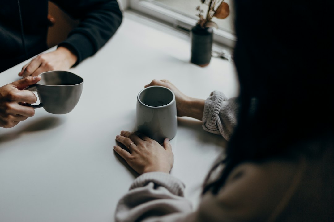 Due persone al tavolo tengono in mano tazze durante una conversazione; immagine illustrativa dell'ascolto senza fretta. Foto: Priscilla Du Preez / Unsplash.